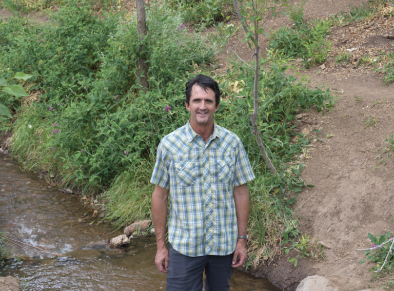 Jesse Roach smiles at the camera while standing in a Santa Fe river surrounded by plants